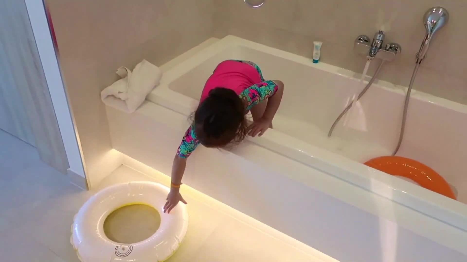 Watch this adorable little girl having a blast in the bathroom! She's reaching out for her white swim ring with colorful sleeves, and there's an orange one floating in the tub too. Bath time turns into a fun adventure with pool toys! 💦😊🎈