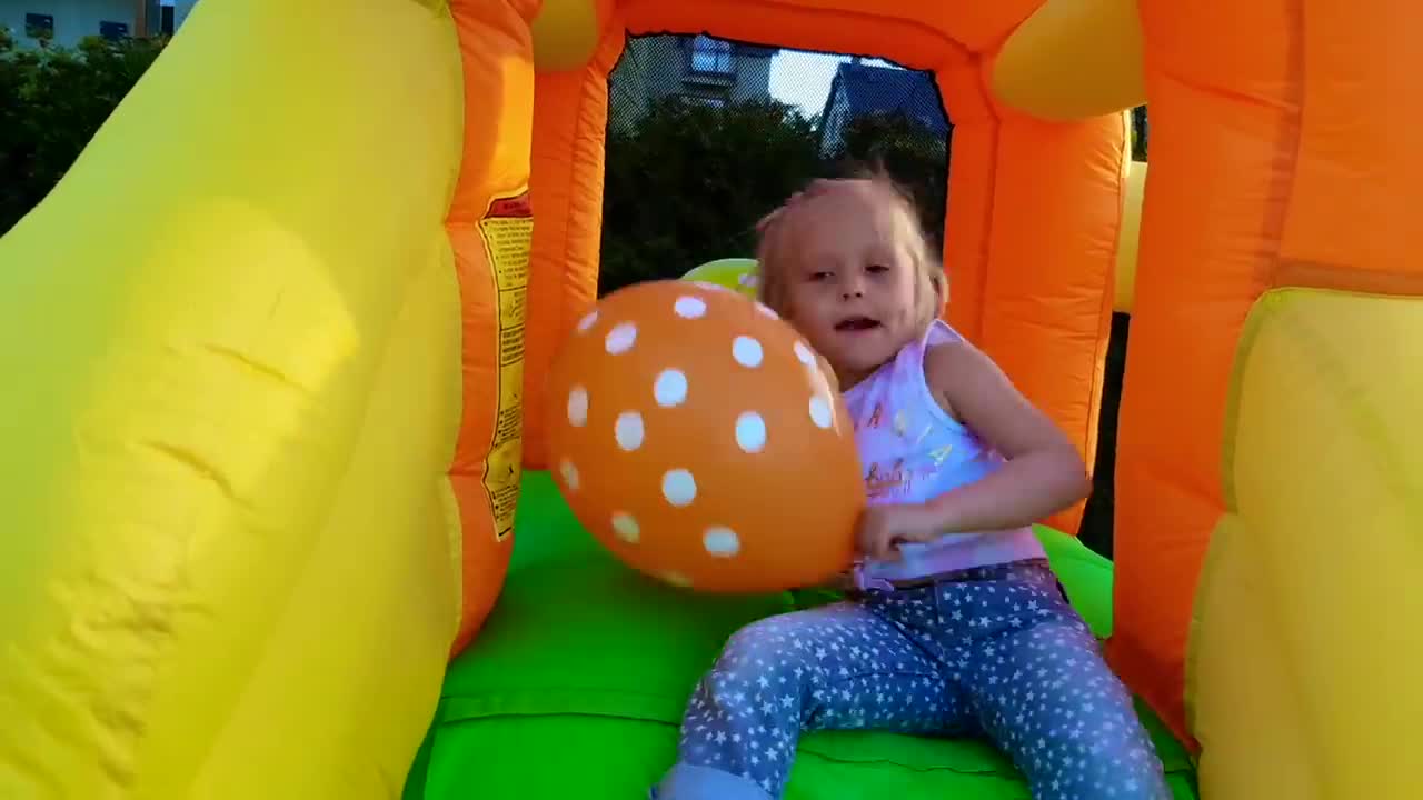 A happy little girl is having fun in a colorful inflatable bounce castle! She’s holding a playful polka-dot balloon, bouncing around and laughing, enjoying a joyful outdoor playtime. 🌈✨
