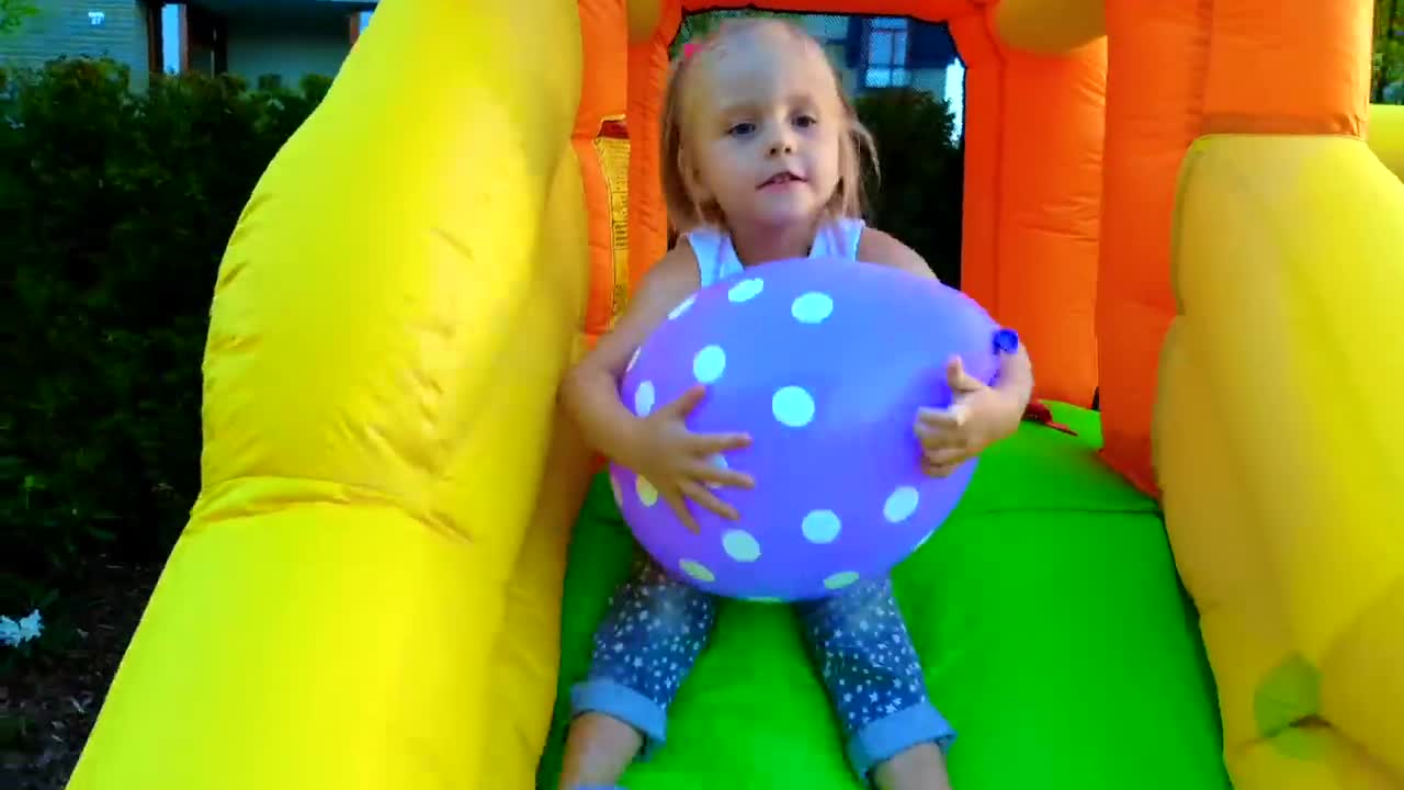 A cute little girl enjoys playing in a colorful bouncy castle! She's holding a fun purple polka dot ball and looks so happy, smiling and having a great time outdoors. It's a sweet moment of carefree childhood joy! 🎉