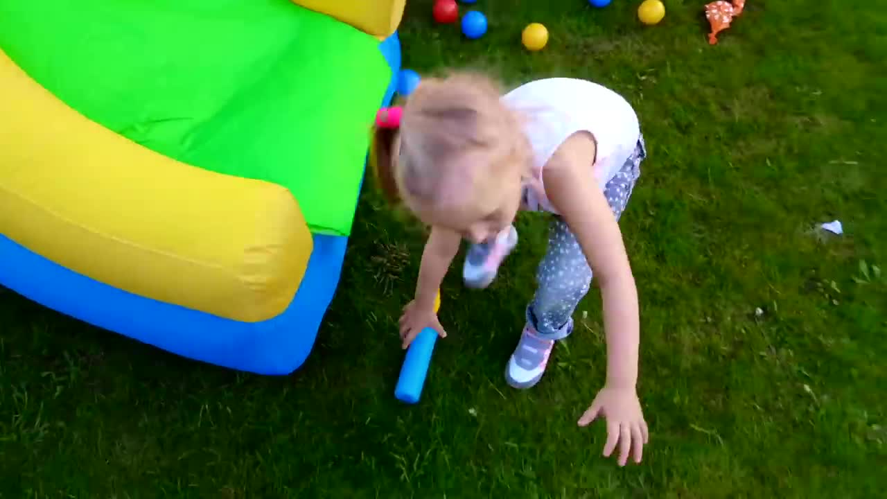 A little girl is having a wonderful time playing outside! She’s near a colorful, inflatable bouncy castle, holding a blue foam stick, and there are bright, playful balls (red, yellow, blue) scattered on the green grass around her. It looks like a day full of giggles and adventure as she enjoys her fun toys! 🎈✨