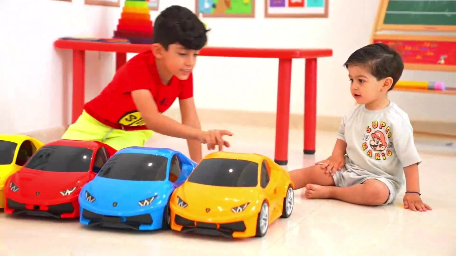 Two little boys are having fun playing with colorful toy cars in a bright room! One boy (wearing a red shirt) is pointing at the yellow car, while the other (in a Mario t-shirt) sits nearby, watching and smiling. They seem to be enjoying a playful time together with their cool toy cars! 😊✨