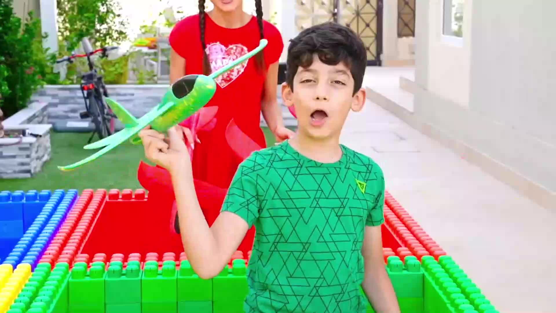 Look! A big, colorful block fort made of red, blue, green, and yellow bricks! A little boy is holding a cool green toy plane, and his friend in a red dress is right there with him. They're playing outside, having so much fun building and flying the plane ๐ #ToyFun #BuildingBlocks #PlayTime #KidsPlay