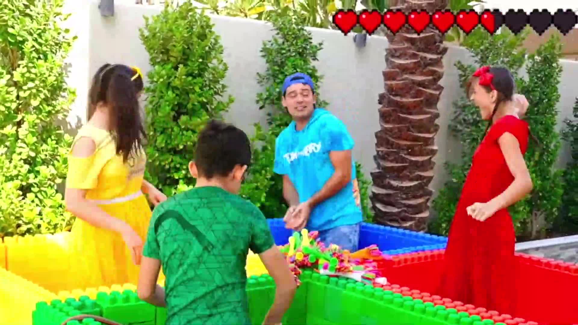 A fun outdoor scene with kids and an adult playing in a colorful building block area! They're surrounded by green plants, and there are playful toys nearby. The vibe is full of joy and creativity, with heart icons 💕 adding to the happy moment!