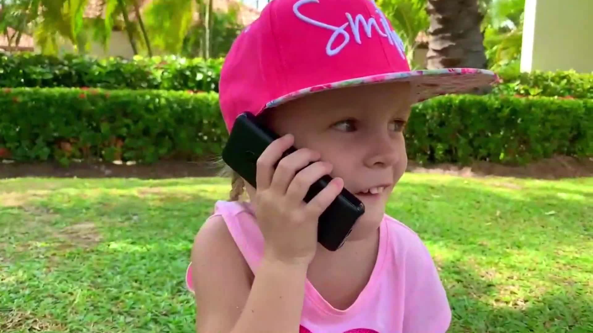 A sweet little girl with a pink 'Smile' hat and a matching top is having a phone call in a sunny, green park. She looks so happy and curious, enjoying the fresh air and beautiful surroundings! 😊🌴