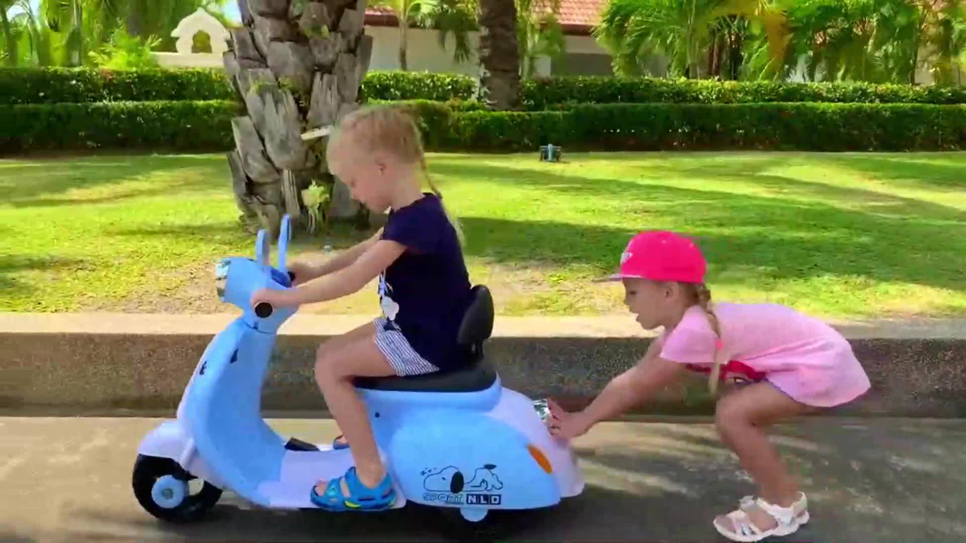 Cute little girls are having the best time! One is zooming on a blue toy scooter, and the other is pushing it, giggling and playing together in the sunny garden. Fresh air, green grass, and happy smiles—pure fun for kids! 🌞🏡