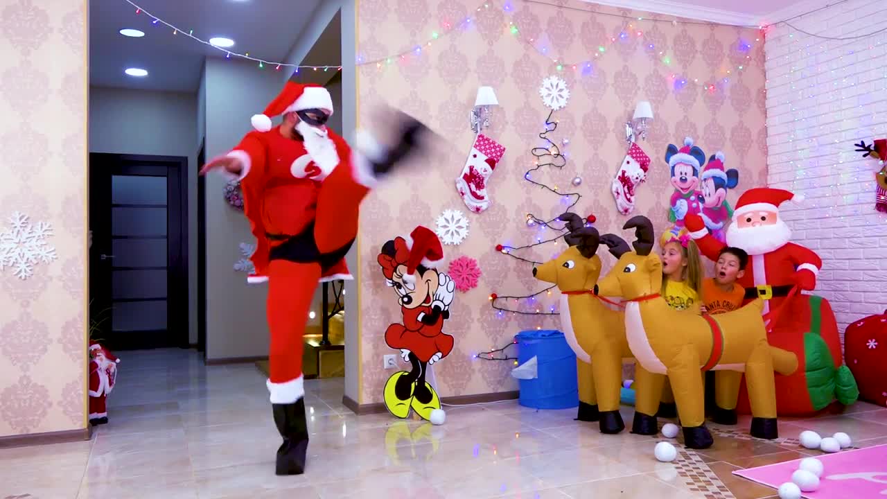 A lively Santa Claus in a red suit is dancing and jumping in a festive room! Colorful Christmas lights, Mickey Mouse decor, and reindeer inflatables make the scene magical. Two kids giggle and watch happilyโfull of Christmas fun and laughter! ๐๐๐ง๐ฆ