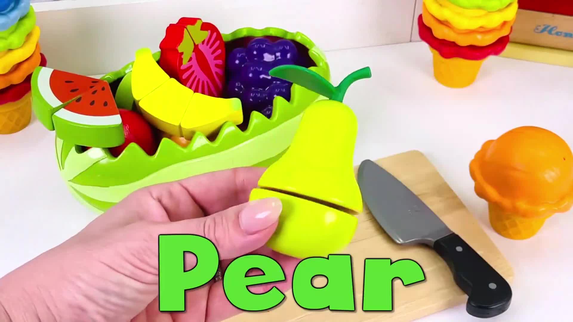 Watch a fun, interactive play session! A hand uses a pretend knife to cut a bright yellow pear toy, with colorful play fruits like bananas, watermelons, and strawberries in a green bowl nearby. Great for little learners to explore fruit names and cutting play! ๐๐