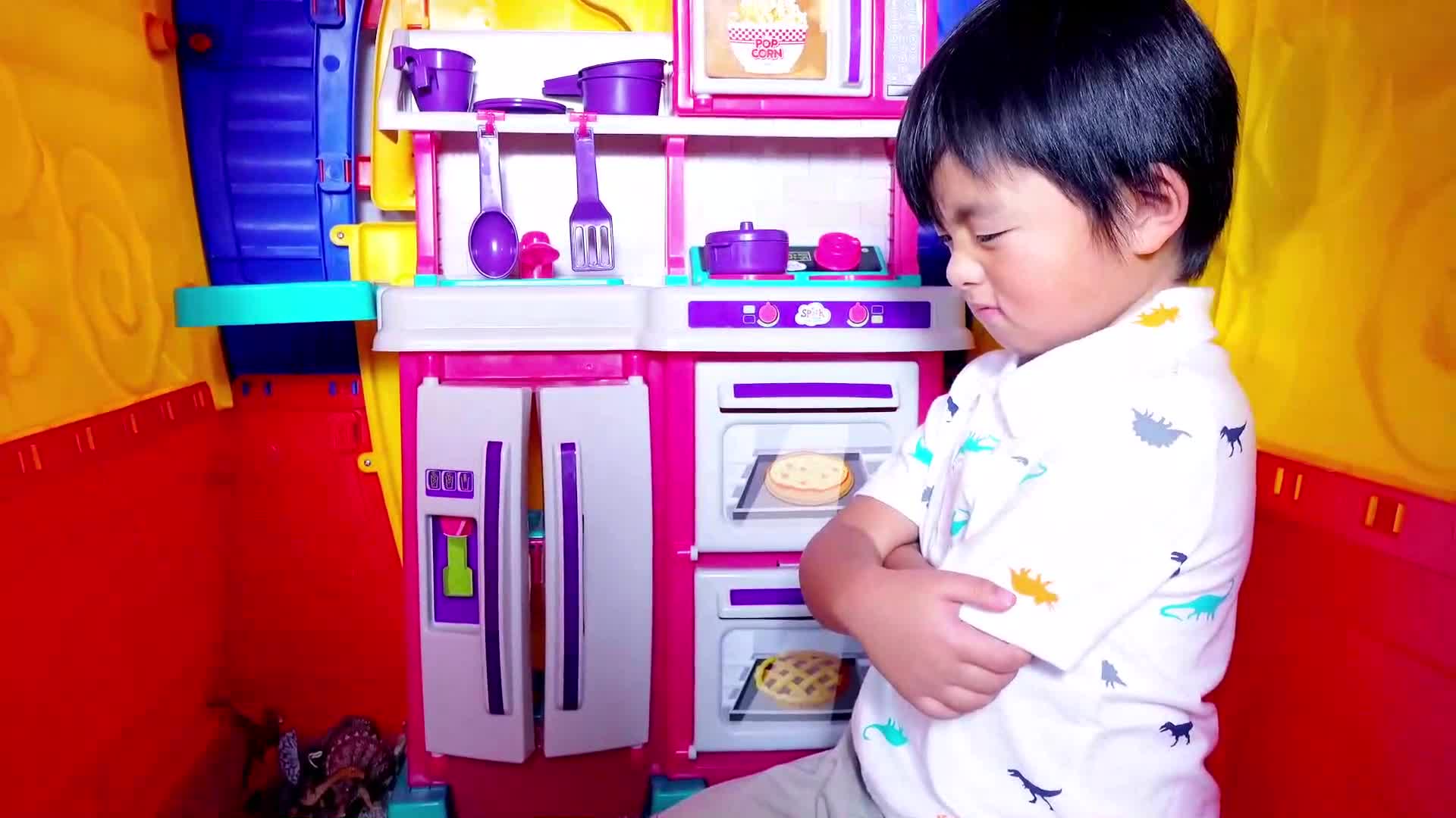 A cute little boy wearing a dinosaur-patterned shirt stands next to his colorful toy kitchen, looking upset with crossed arms! His pink and purple toy kitchen has a fridge, stove, and fun pretend food like cookies and pies baking in the oven. What could be making him mad? Watch to find out! ๐