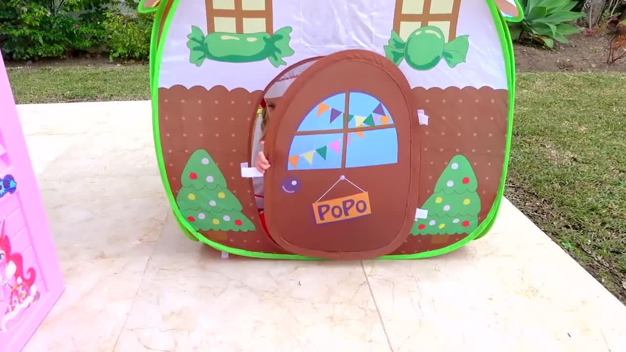 A cute little girl is having fun playing inside a colorful candy-themed play tent! The tent is decorated with big green candies, Christmas trees with colorful ornaments, and a "PoPo" sign on the door. She’s peeking out the door, probably exploring her new playhouse. Outside, there’s a pink toy house nearby, making this a super fun outdoor play spot for kids! 👧🎄✨