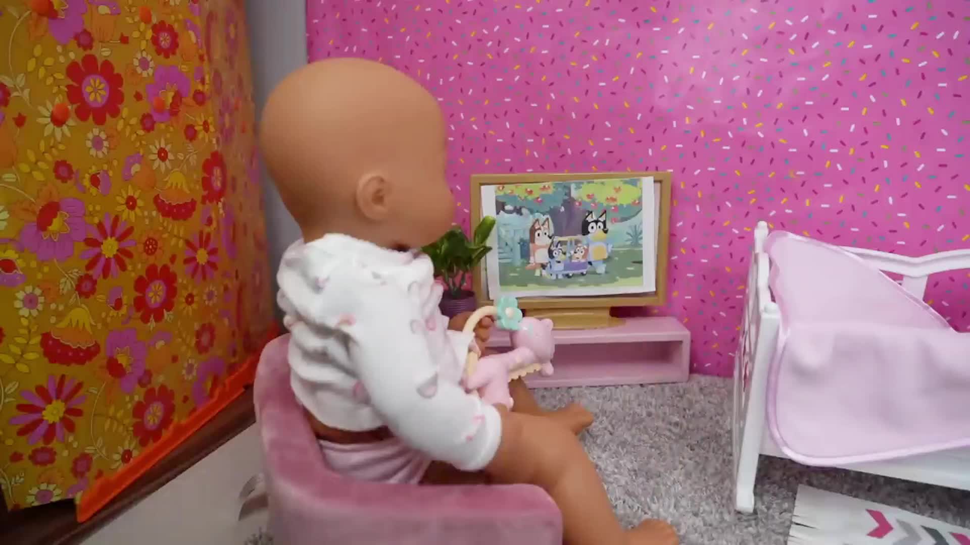 A cute baby doll sits in her cozy pink chair, holding a soft toy, and watches Bluey and friends on the tiny TV screen! 💕 The room has pretty sprinkled pink walls, a little bed, and a colorful floral corner—perfect for a sweet, playful scene! 🥰
