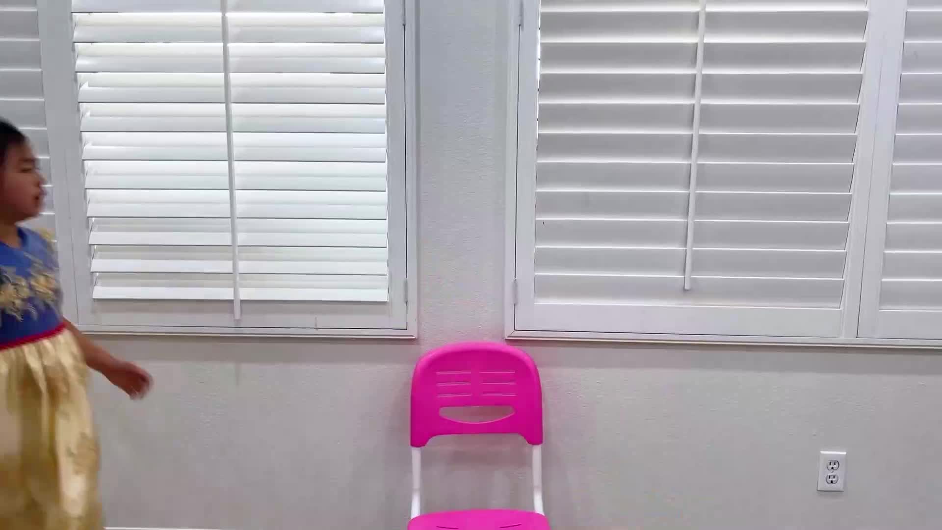 A cute little girl wearing a pretty blue and gold dress is moving around a bright room with white shutters and a pink plastic chair nearby! 👧🎈🌈